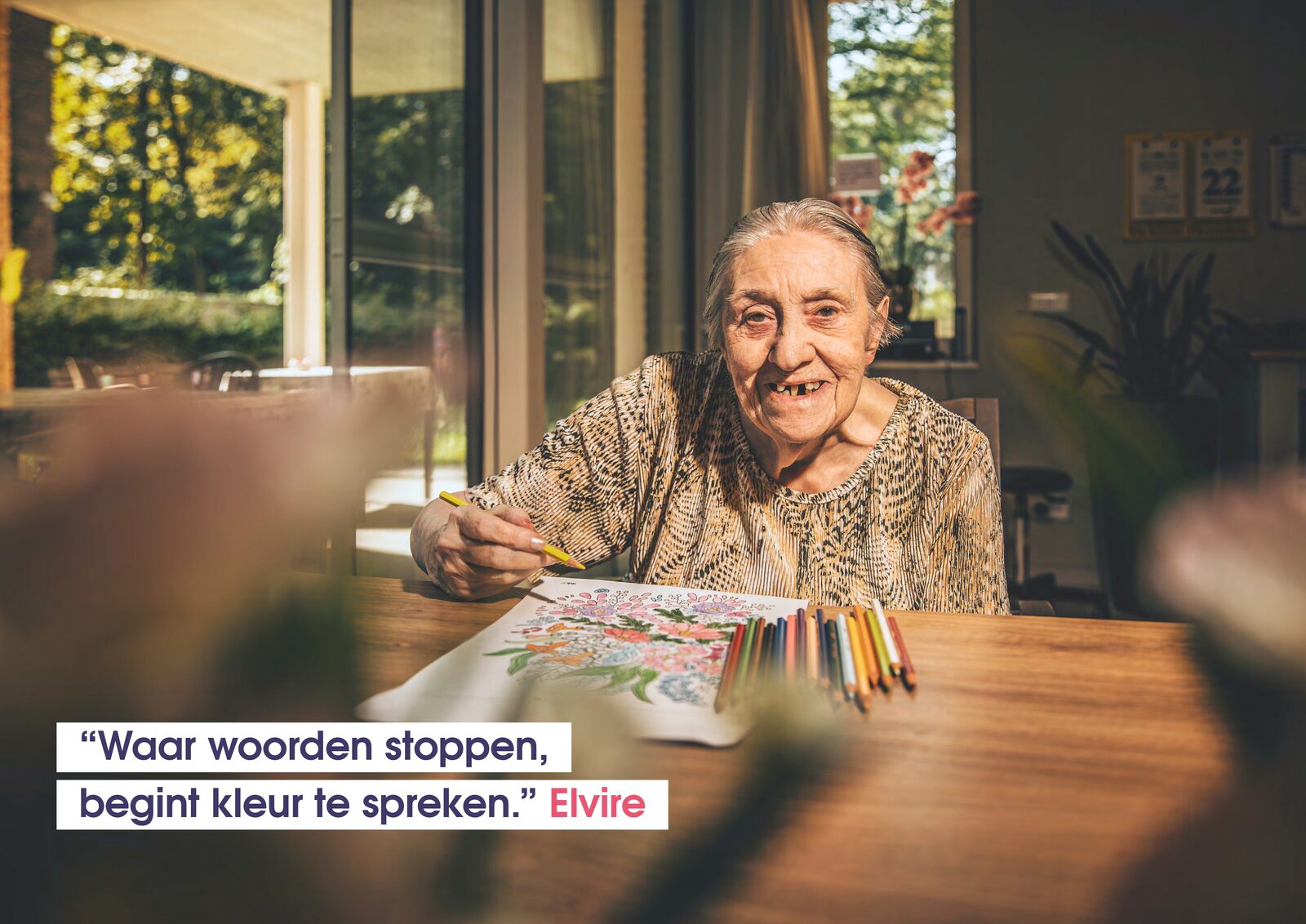 Elvire kleurt een bloemmotief aan een tafel met kleurpotloden, bij een raam met zicht op de tuin.